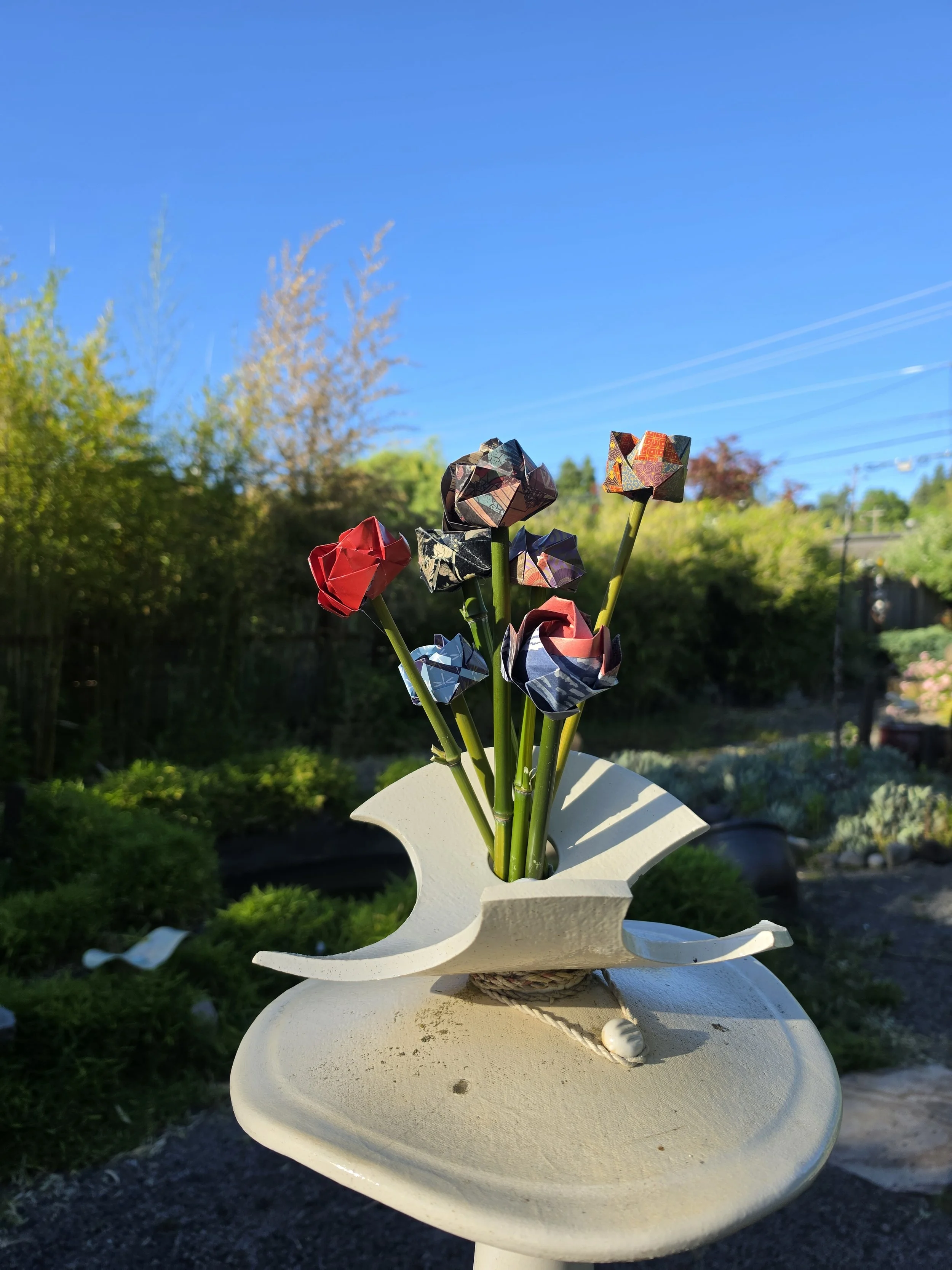 A decorative sculpture of origami flowers made from patterned paper, mounted on green stems, displayed on a white pedestal outdoors under a clear blue sky with trees in the background.