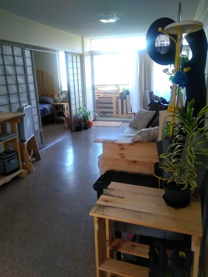 A cozy living room with a wooden sofa, potted plants, a telescopic viewer, and a sliding glass door leading to a balcony with wooden pallets.