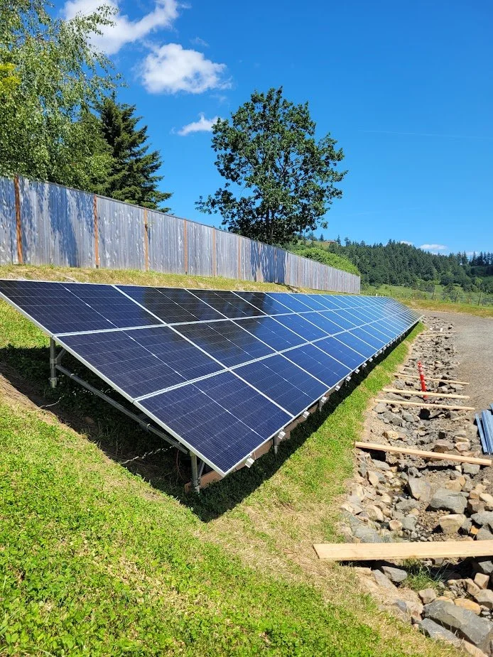 A row of solar panels installed on a grassy hill with a wooden fence and trees in the background, under a clear blue sky.