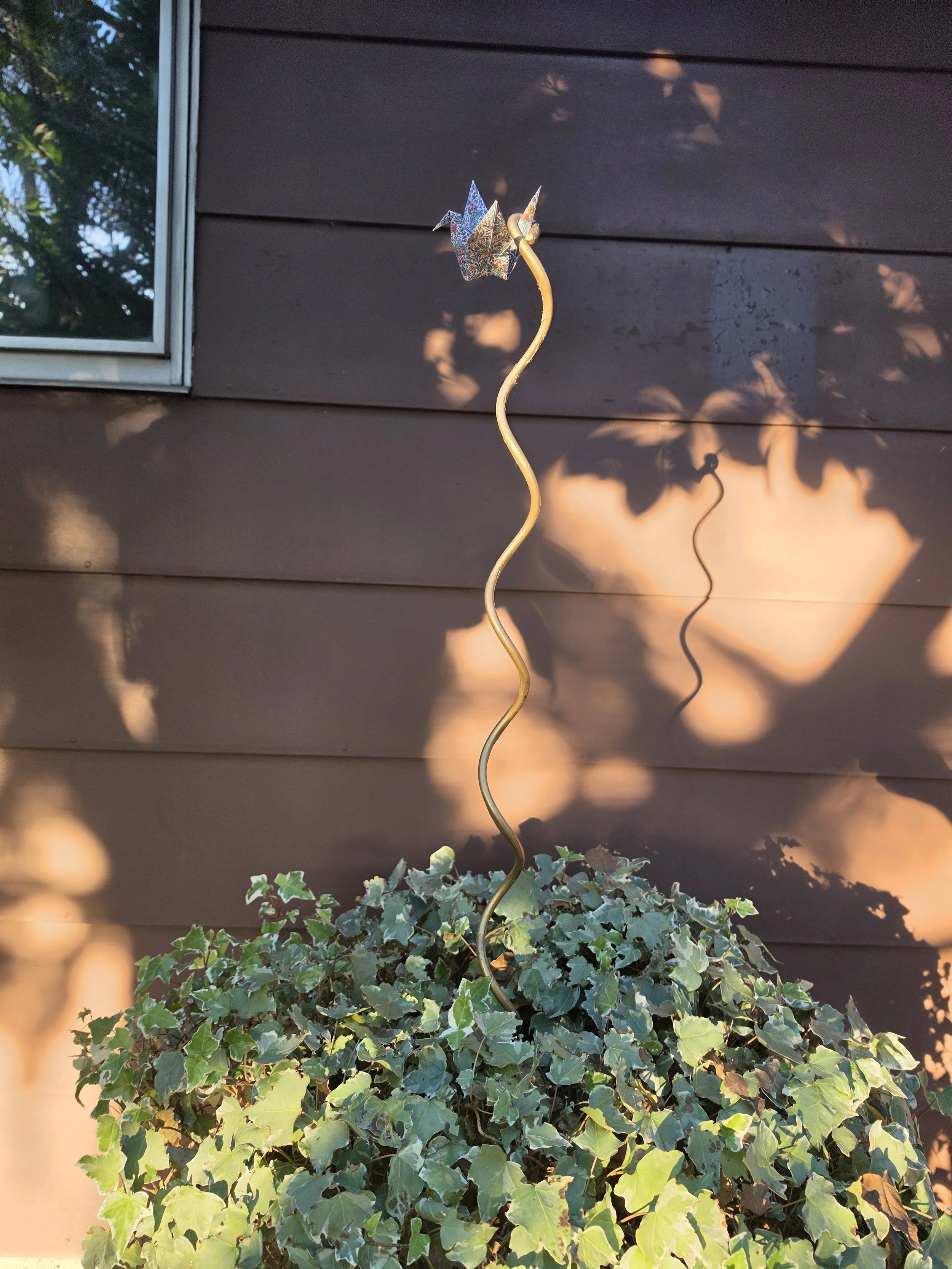 A decorative metal garden stake shaped like a paper origami crane with a curly stem, placed among green ivy plants against a dark brown wooden wall, with sunlight and shadows creating a warm glow.