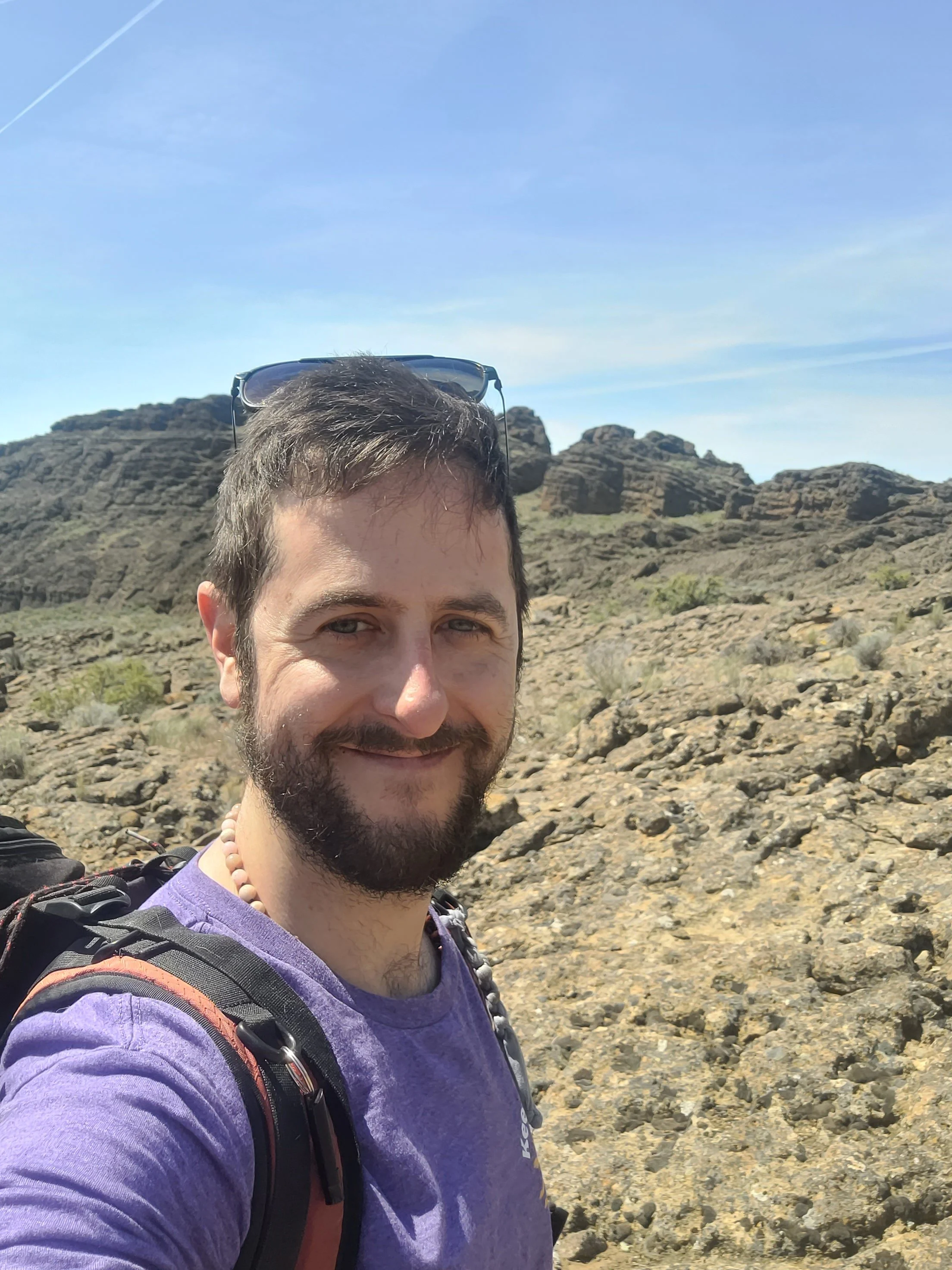 A man with short brown hair and a beard smiling outdoors in a rocky desert landscape with blue sky and some clouds.