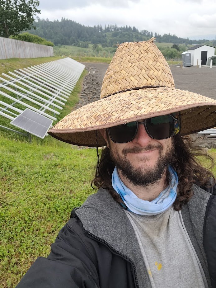 Person wearing a wide-brimmed straw hat and sunglasses, smiling outdoors near solar panels on a cloudy day.
