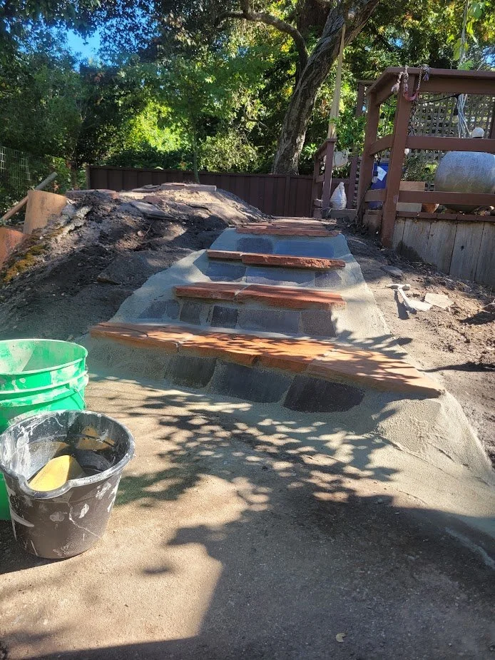 Construction scene of outdoor staircase with bricks and concrete, surrounded by trees and outdoor tools.