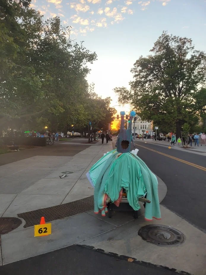 A large, colorful snail costume draped over a vehicle on a city sidewalk at sunset, with trees, buildings, and pedestrians in the background.