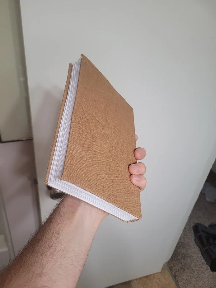A person holding a large book with a plain, brown cover against a beige wall.