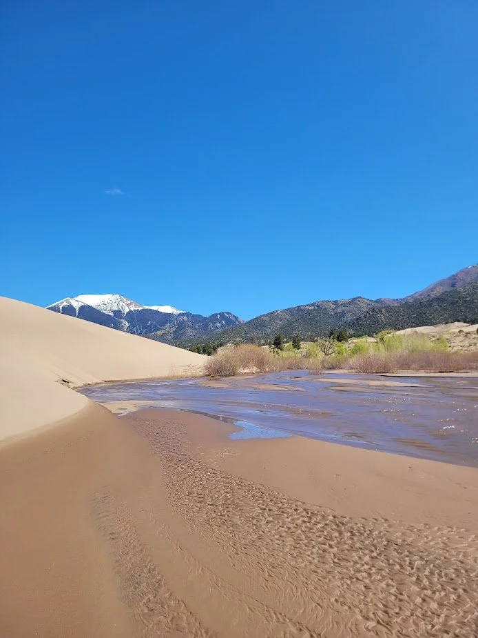 Desert landscape with a sand dune, a river flowing through the sand, mountains with snow in the background, and a bright blue sky.