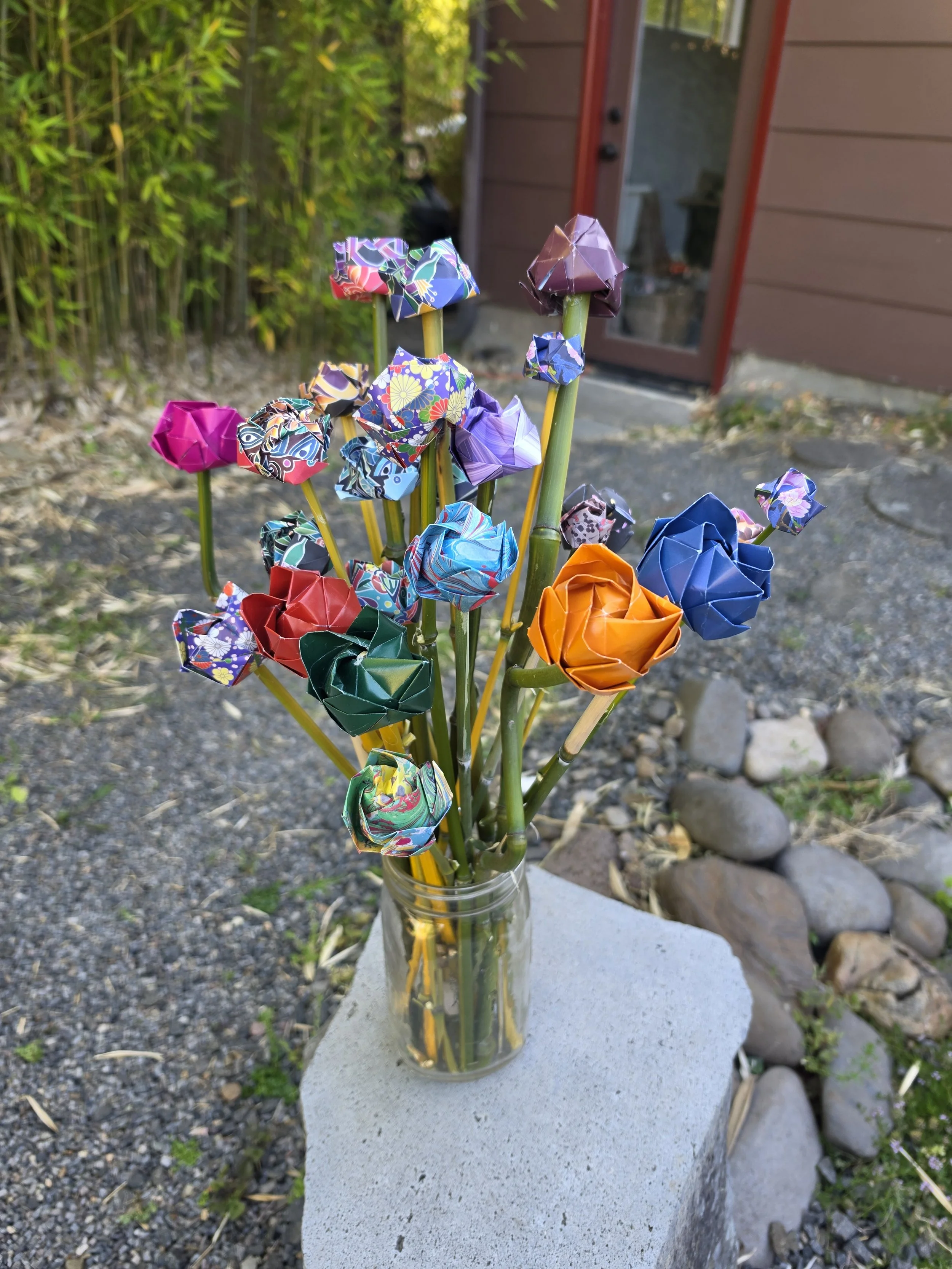 A glass jar with colorful origami paper flowers on yellow stems, placed on a concrete surface outdoors near rocks and a brown building with a glass door in the background.