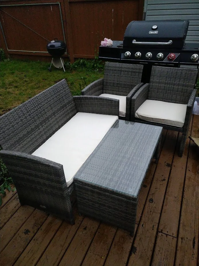 Outdoor patio furniture set on a wooden deck, including a gray wicker sofa with white cushions, two matching armchairs, and a rectangular glass-topped coffee table. In the background, a black grill with four burners, and a small charcoal grill, with a pink flower decoration visible on the grill.