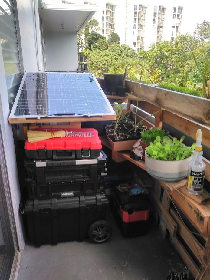 Small urban balcony with solar panel, tools, plants, and gardening supplies in a city apartment setting.
