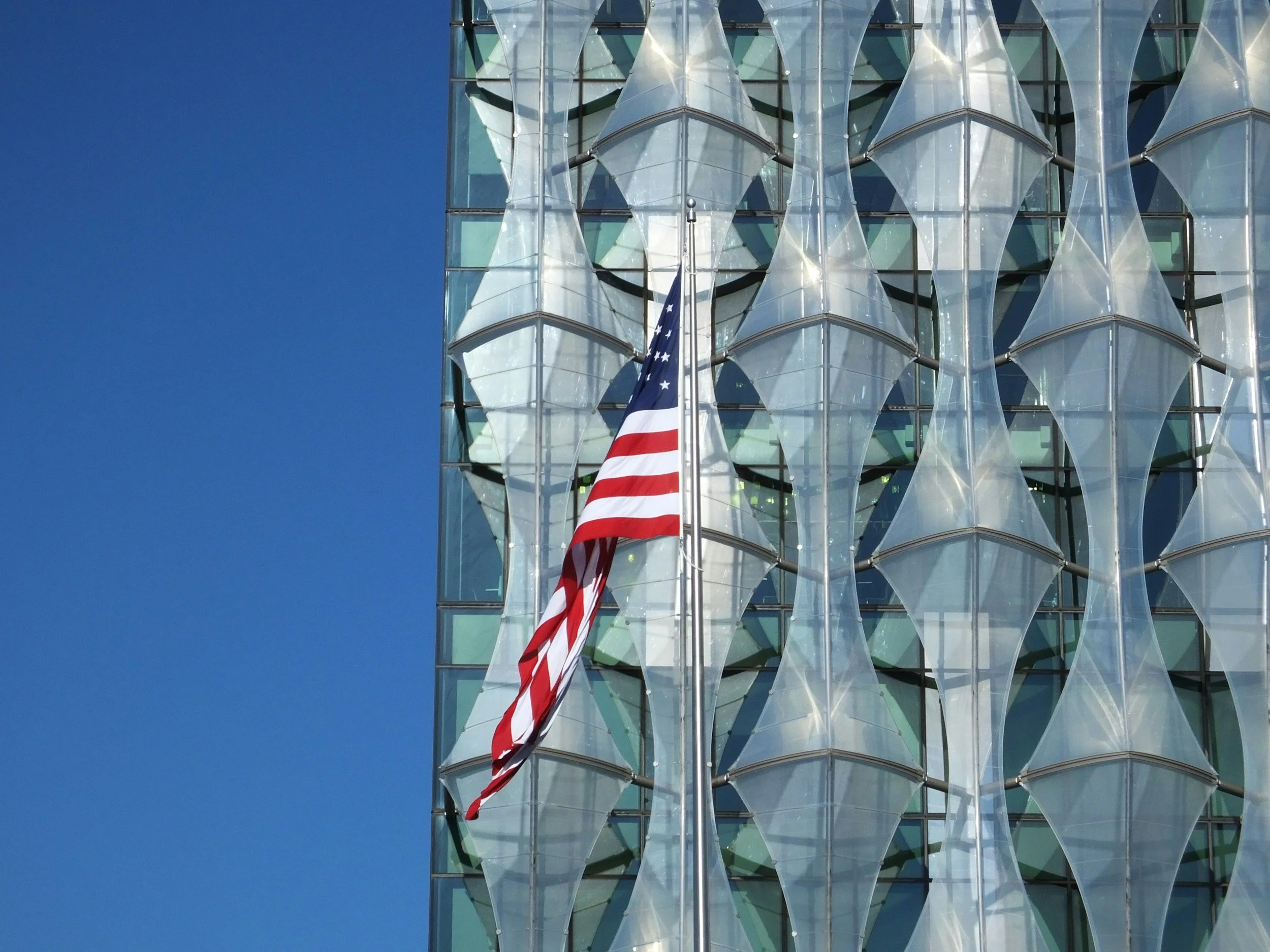 Close-up of a modern building with a glass facade featuring wavy, reflective panels and an American flag flying in front.