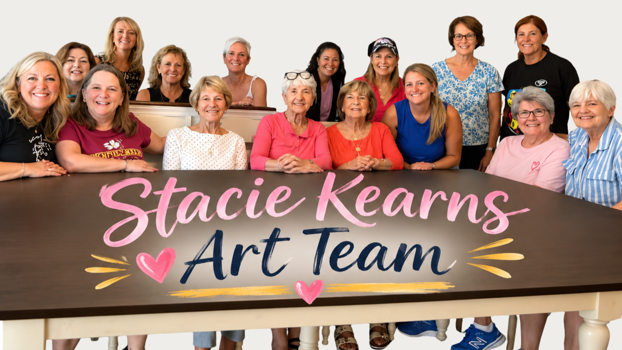 A group of women, mostly middle-aged and older, gathered around a table with a colorful sign that reads, 'Stacie Kearns Art Team,' decorated with pink hearts and yellow accents. The women are smiling and posing for the photo in bright, casual clothing.