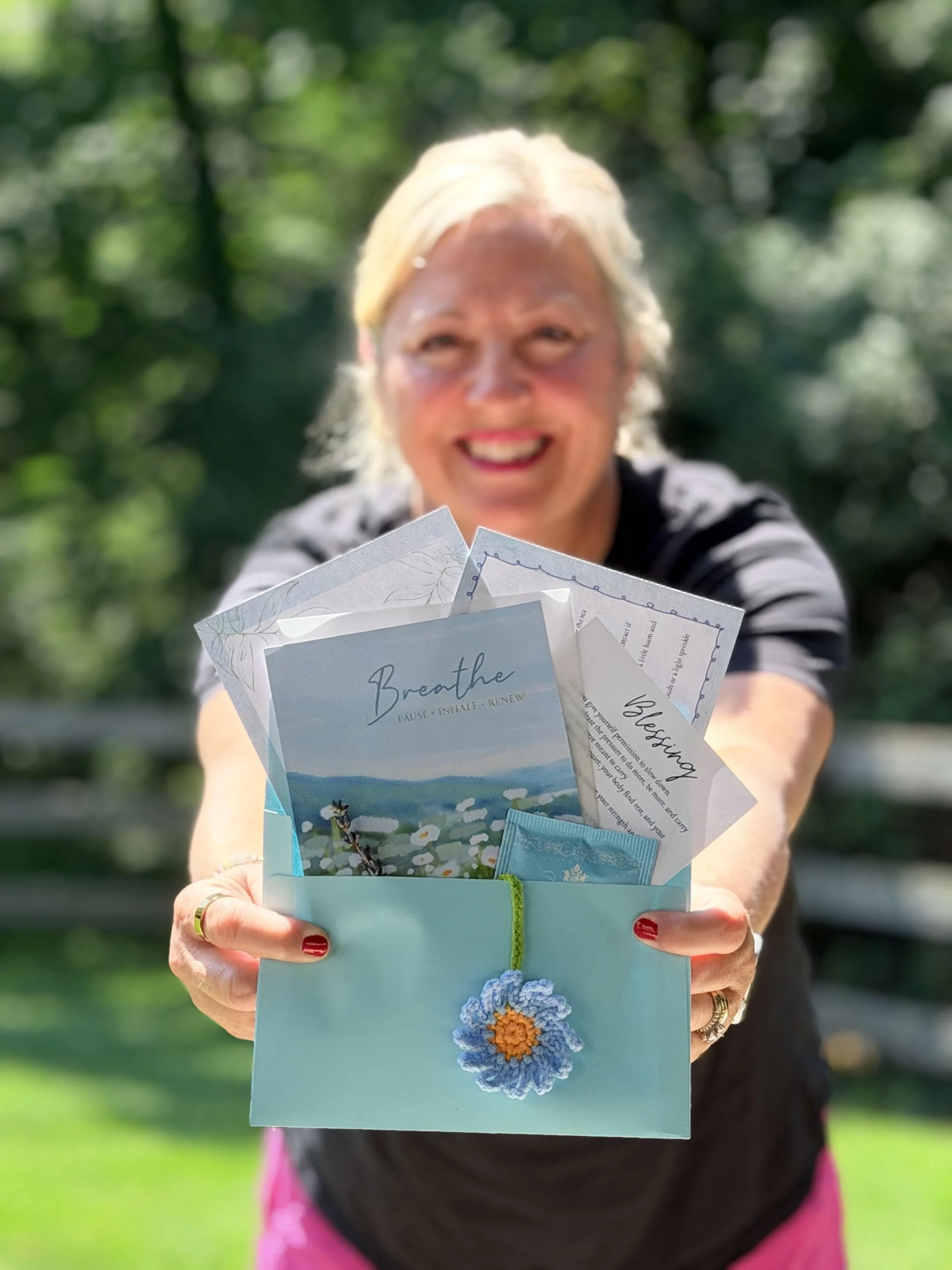 A woman with blonde hair and red painted nails holding a small, decorated envelope with a blue flower and reading material inside, outdoors with blurred green trees in the background.