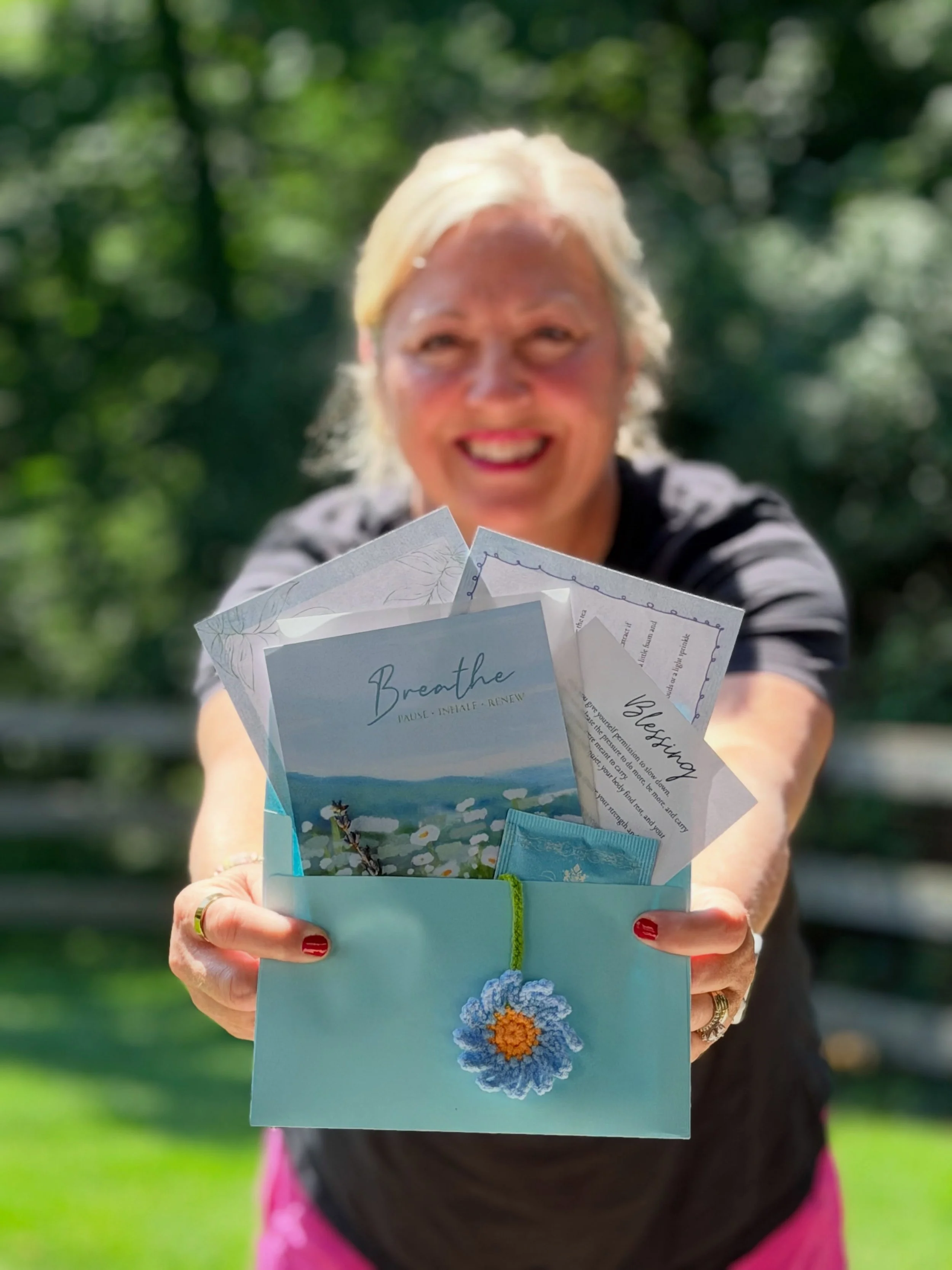 A woman with blonde hair holding a light blue card with a flower decoration, with a background of green trees, outdoors.