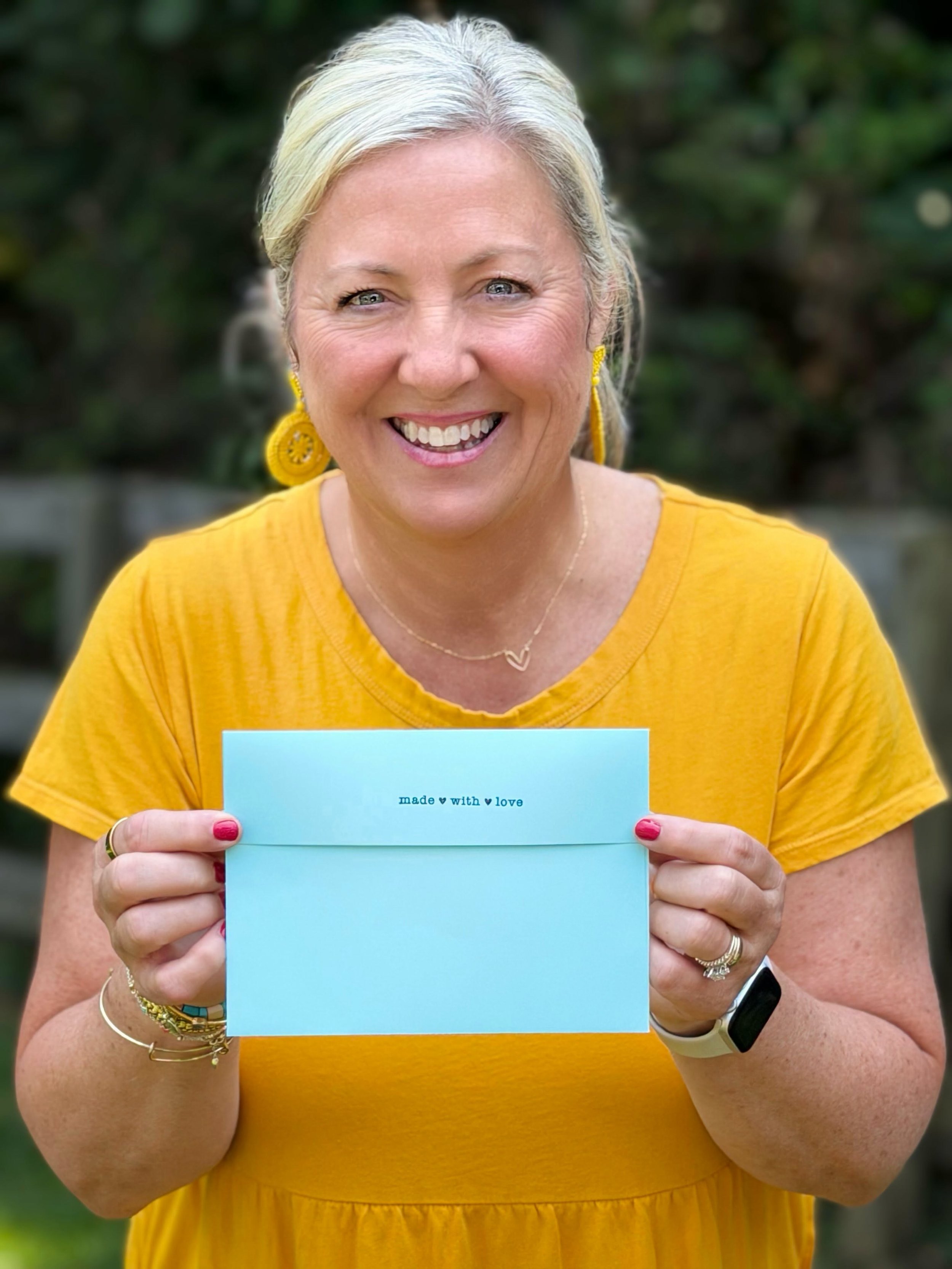 A woman with blonde hair wearing a yellow dress, smiling, holding a light blue card with the words "made with love" printed on it, standing outdoors with green foliage in the background.