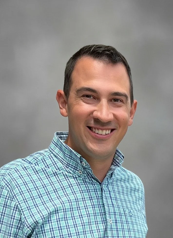A smiling man wearing a checkered button-up shirt standing against a plain gray background.