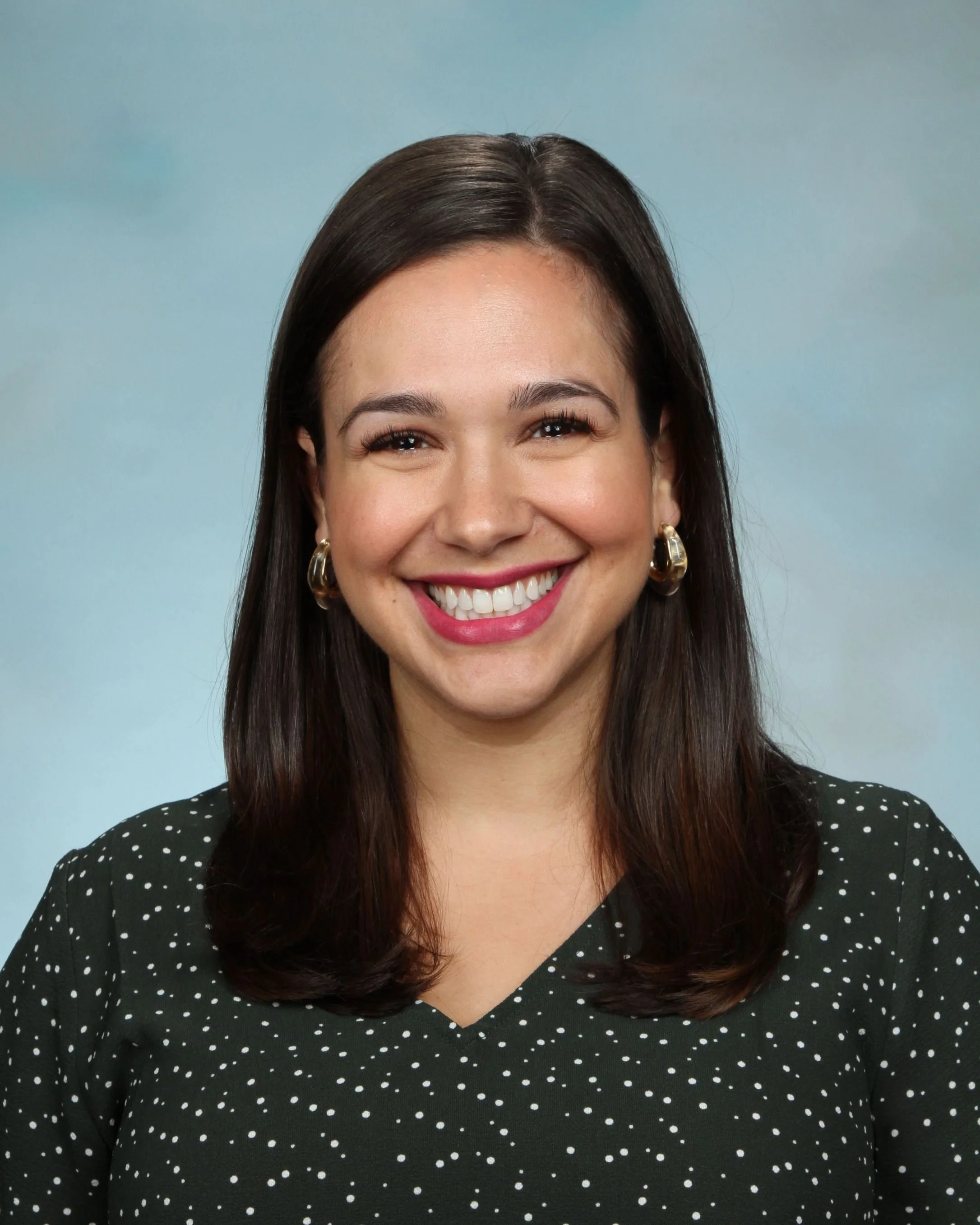 Close-up of a woman with long dark hair, smiling, wearing gold earrings and a black dress with white polka dots, against a light blue background.