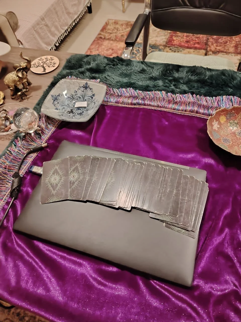 A tarot card deck on a closed laptop, with decorative bowls and figurines on a table covered with a purple cloth, in a cozy room.