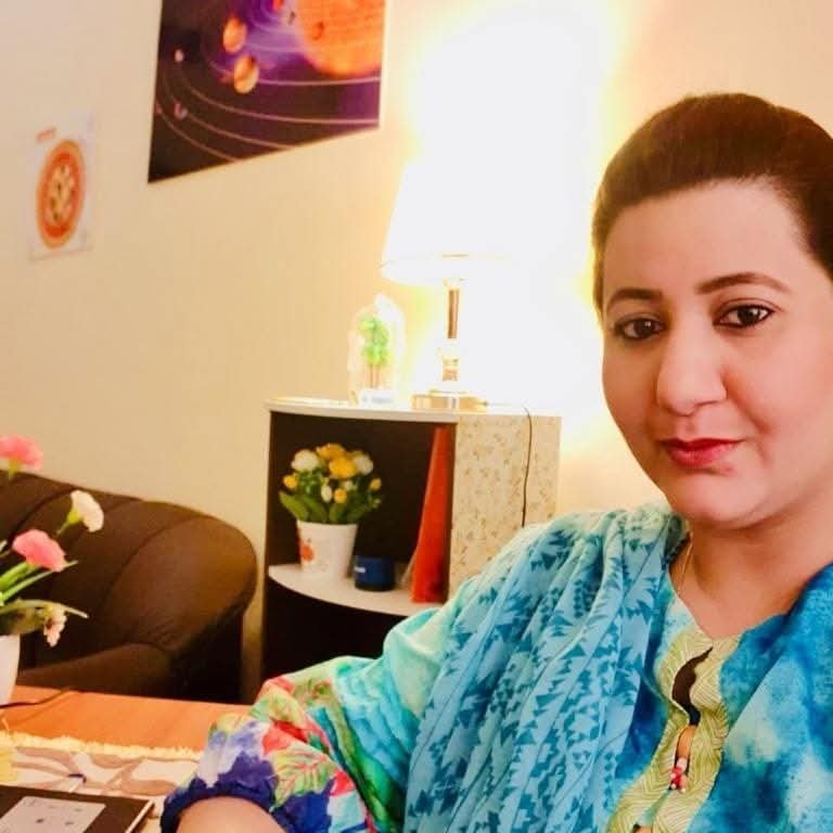 A woman with dark hair tied back, wearing a colorful blue and green outfit, taking a selfie in a cozy living room with a beige shelf, a lamp, and a potted plant in the background.