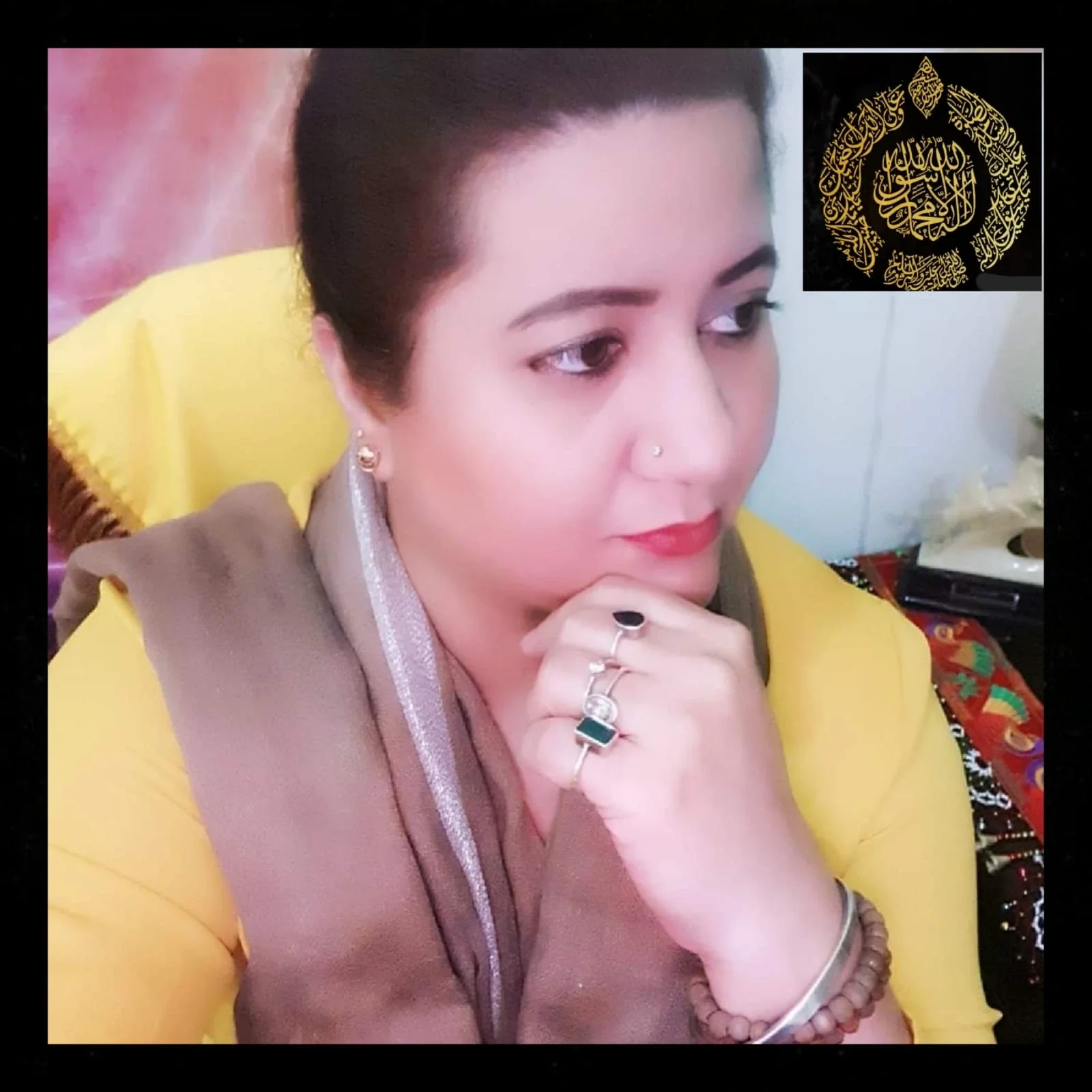 A woman with dark hair and makeup, wearing multiple rings and jewelry, is resting her chin on her hand, looking to the side. She is wearing a yellow and brown outfit with a colorful fabric underneath. There is a decorative black and gold piece with A