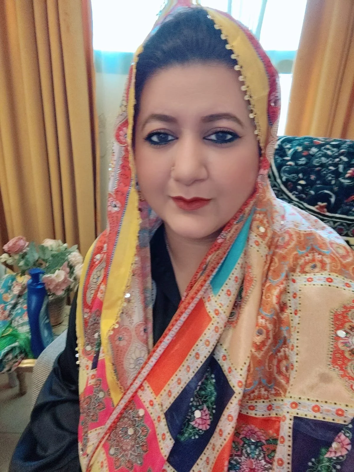 A woman wearing a colorful traditional shawl with intricate patterns, sitting indoors with a floral arrangement and decorative items in the background.