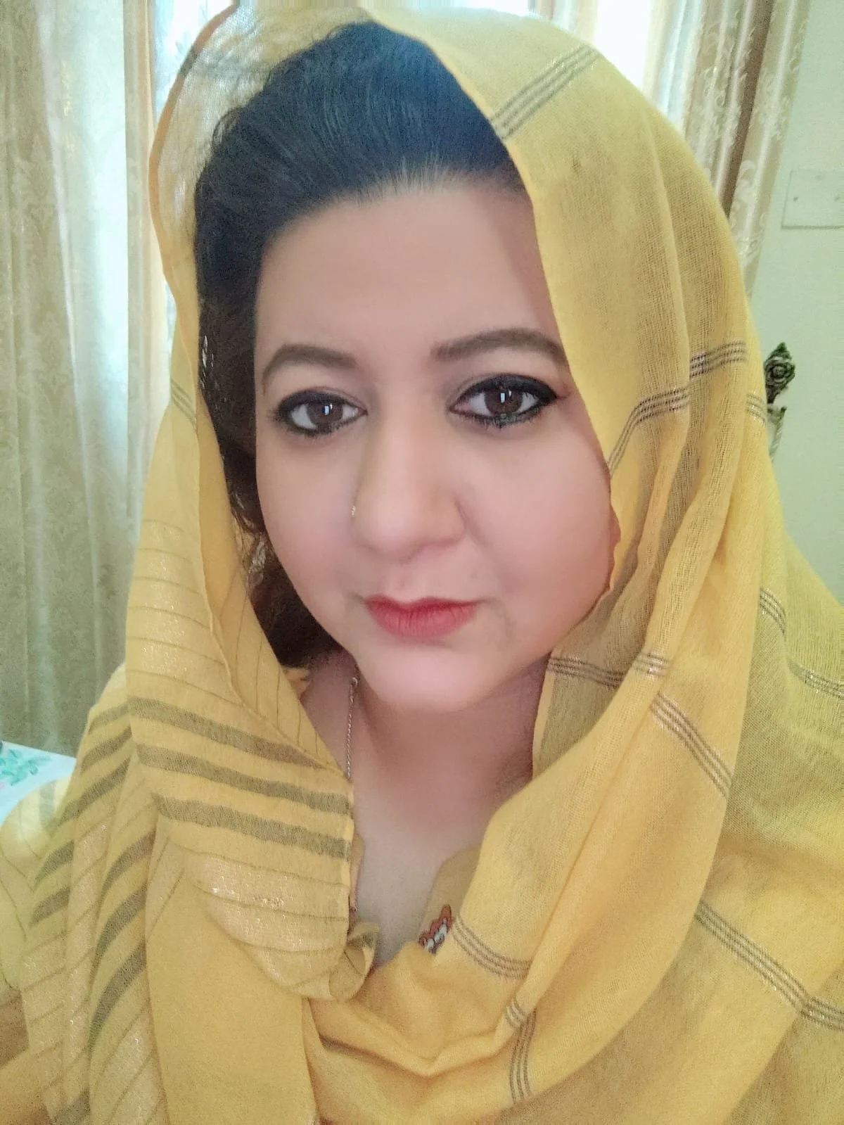 A woman with dark hair and makeup, wearing a yellow headscarf with a striped pattern.