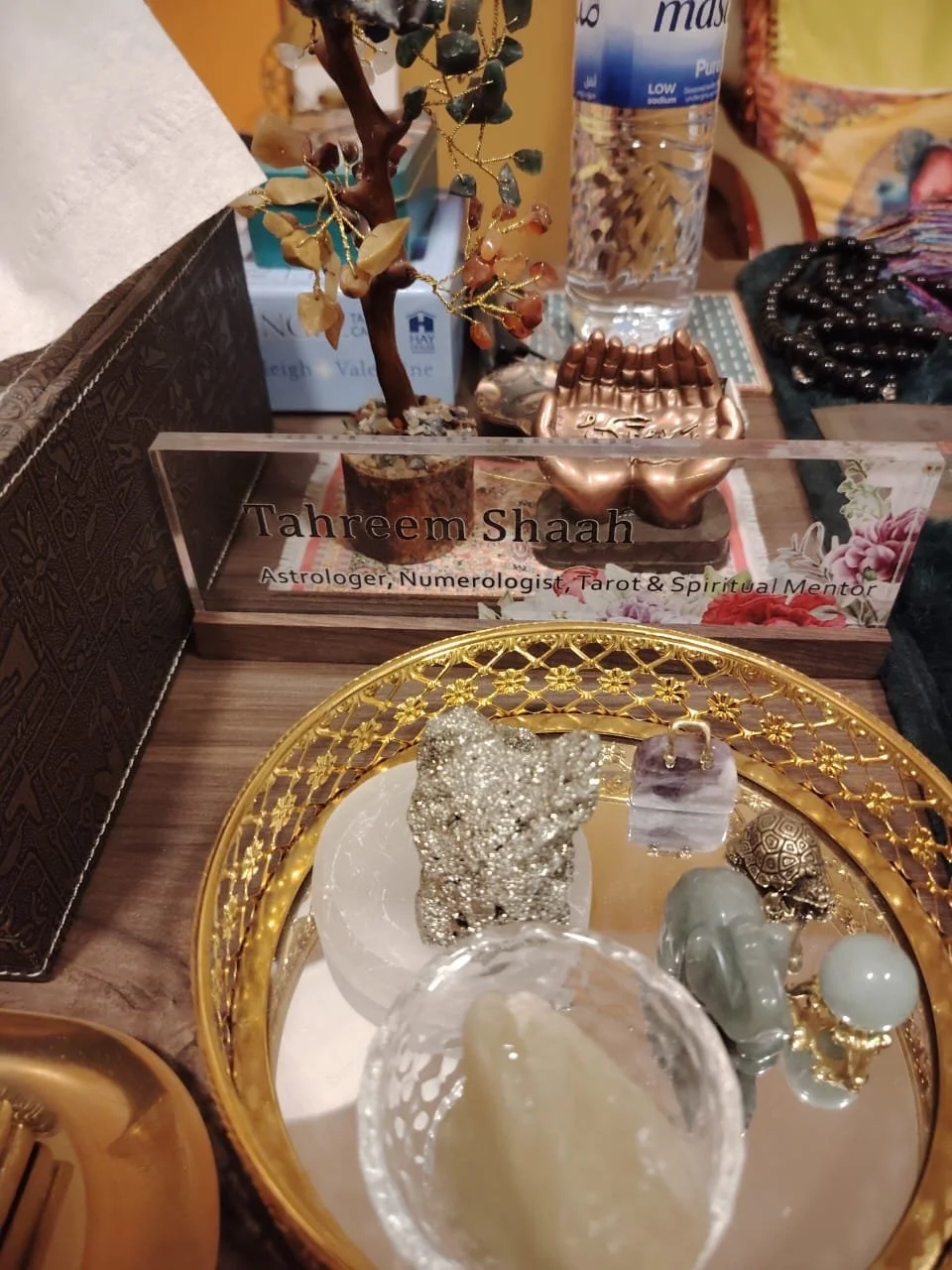 A desk with a nameplate reading 'Tahreem Shaah, Astrologer, Numerologist, Tarot & Spiritual Mentor' behind a small decorative tree and a bottle of water. In the foreground, a gold tray with various crystals and stones.