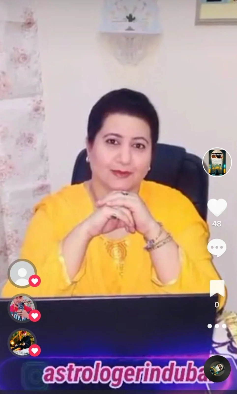 A woman with short dark hair, wearing a yellow outfit, sits at a desk with her hands clasped, smiling at the camera. The background features a white wall with decorative pieces and a laptop in front. The image appears to be a screenshot from a social