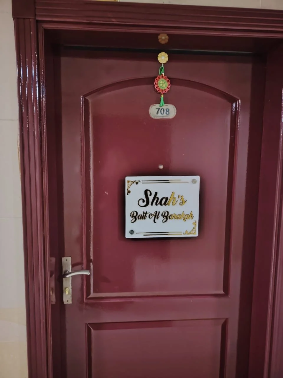 Maroon-colored door with a door handle on the left and a nameplate that reads "Shah's Bait Al Barakah." There is a room number 708 above the door and a hanging decoration with a green tassel.