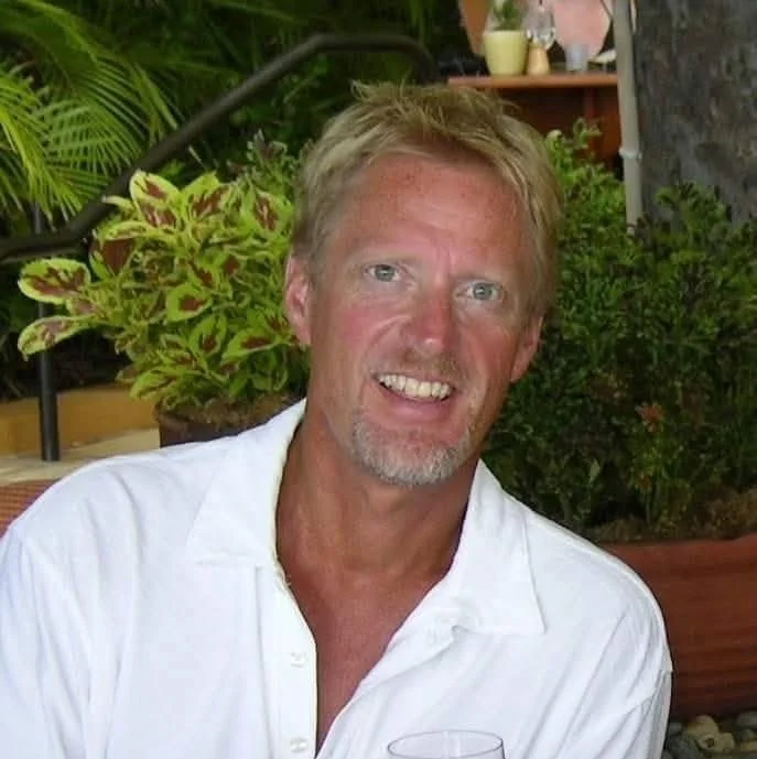 Tim Katzinski, middle-aged man with blonde hair, a short beard, wearing a white shirt, smiling, in a garden setting with plants in the background.