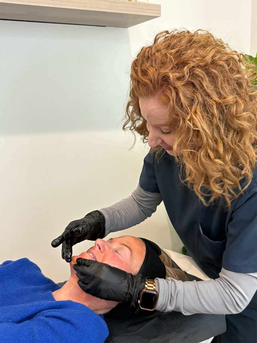 A woman with curly red hair receiving a facial treatment from a professional, both are wearing gloves and face masks in a clinical setting.