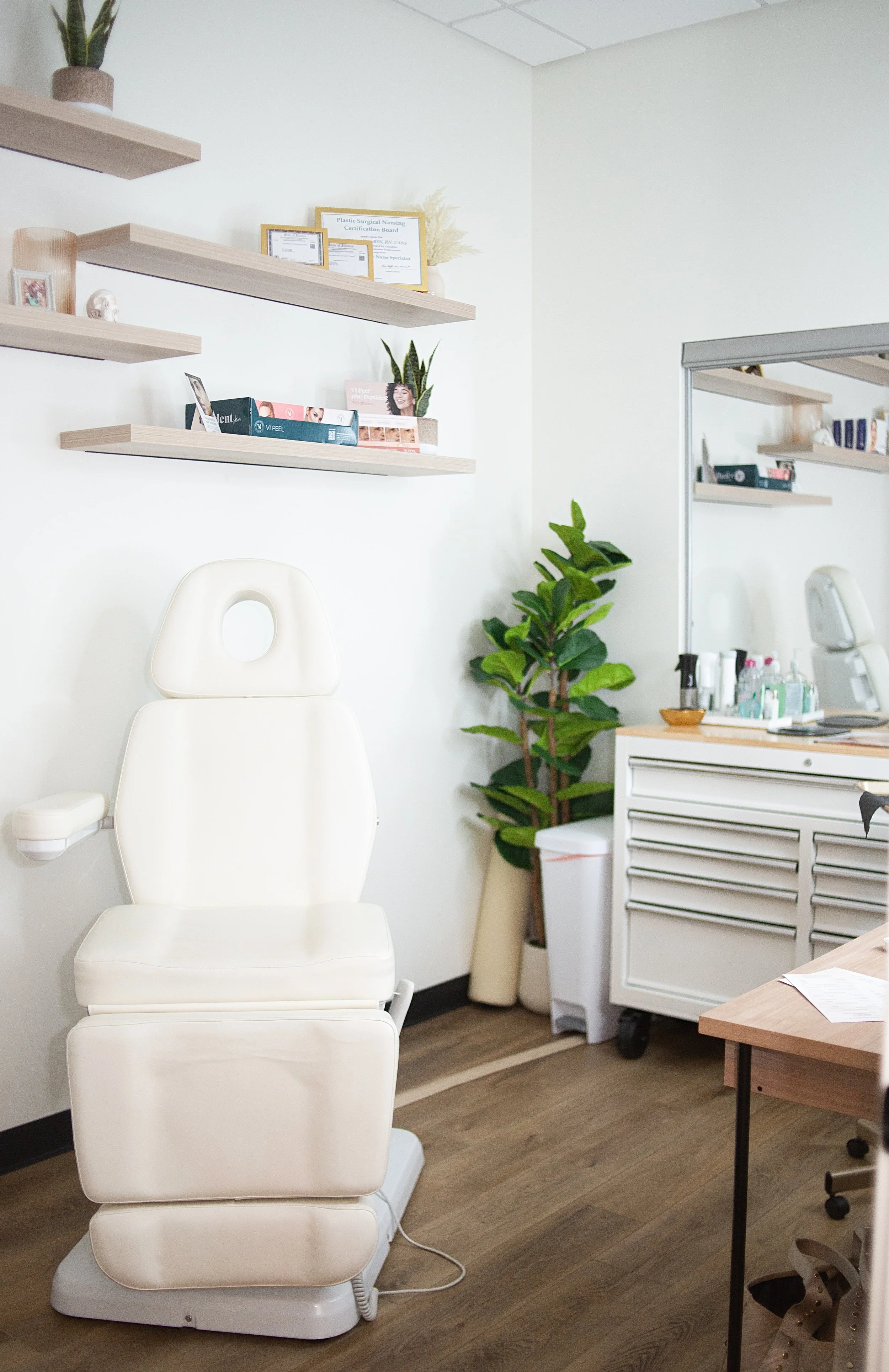 Medical exam room with a white examination chair, shelves with certificates and decor, potted plant, medical supplies on a cart, and a desk with papers and a chair.