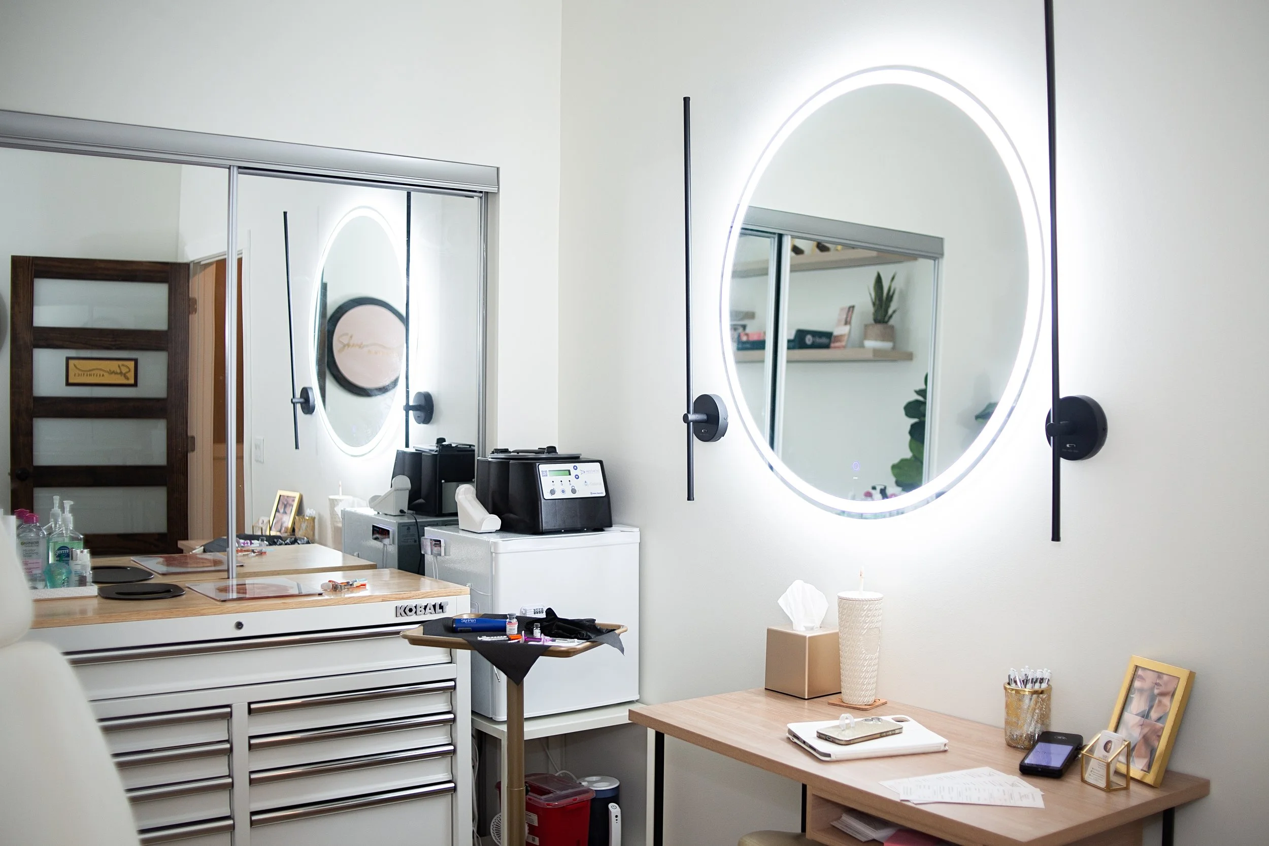 A medical or laboratory office with a white counter, various medical tools, a computer, and two illuminated round mirrors on a wall. There are shelves with books and a plant, and a table with a notepad, a smartphone, and picture frames.