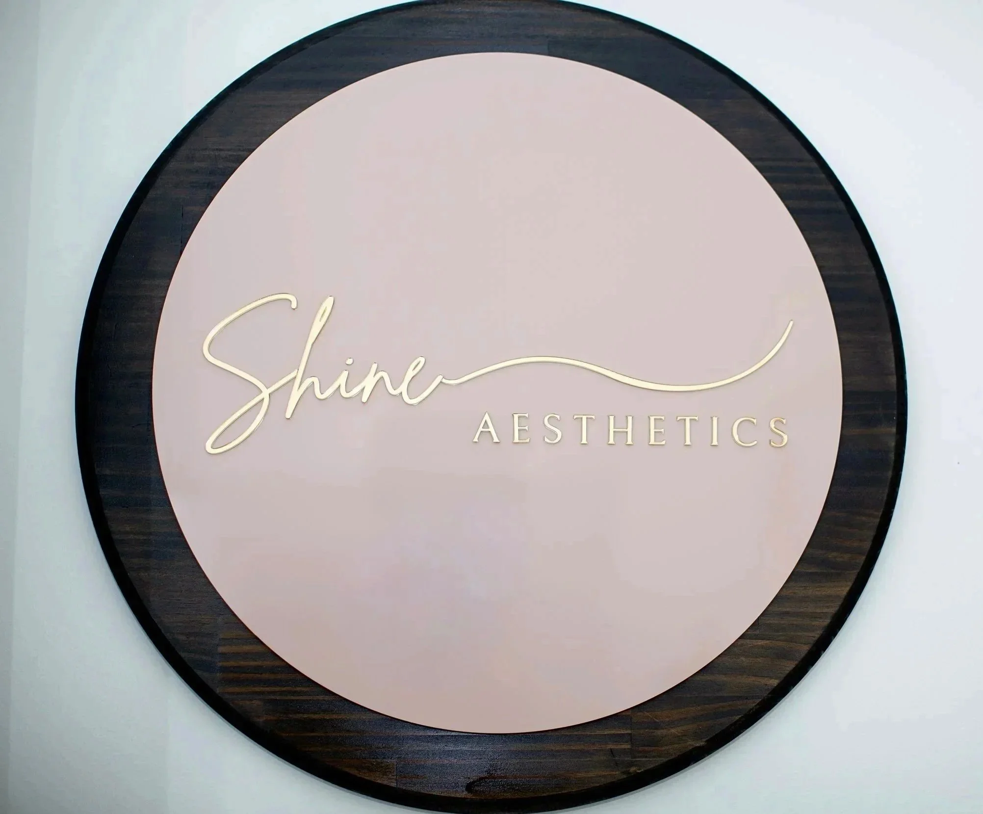 A circular sign with a pink center and black wooden border that reads "Shine Aesthetics" in cursive gold lettering.