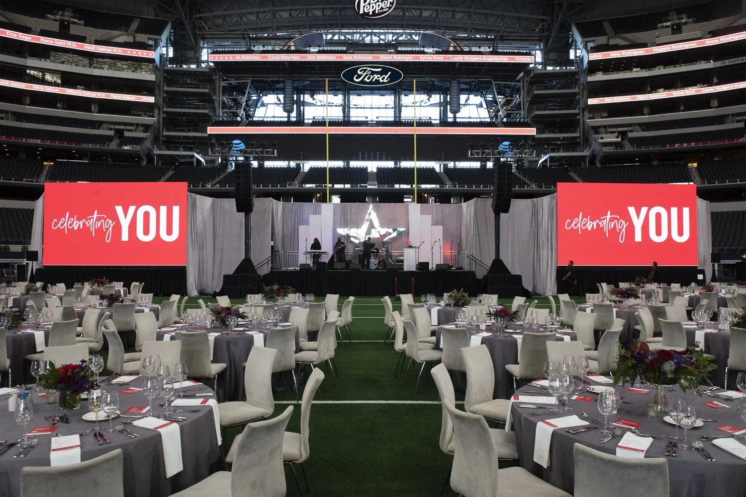 Indoor stadium with a stage, two large screens displaying 'celebrating YOU', and dining tables set with chairs, wine glasses, and floral centerpieces, prepared for an event.