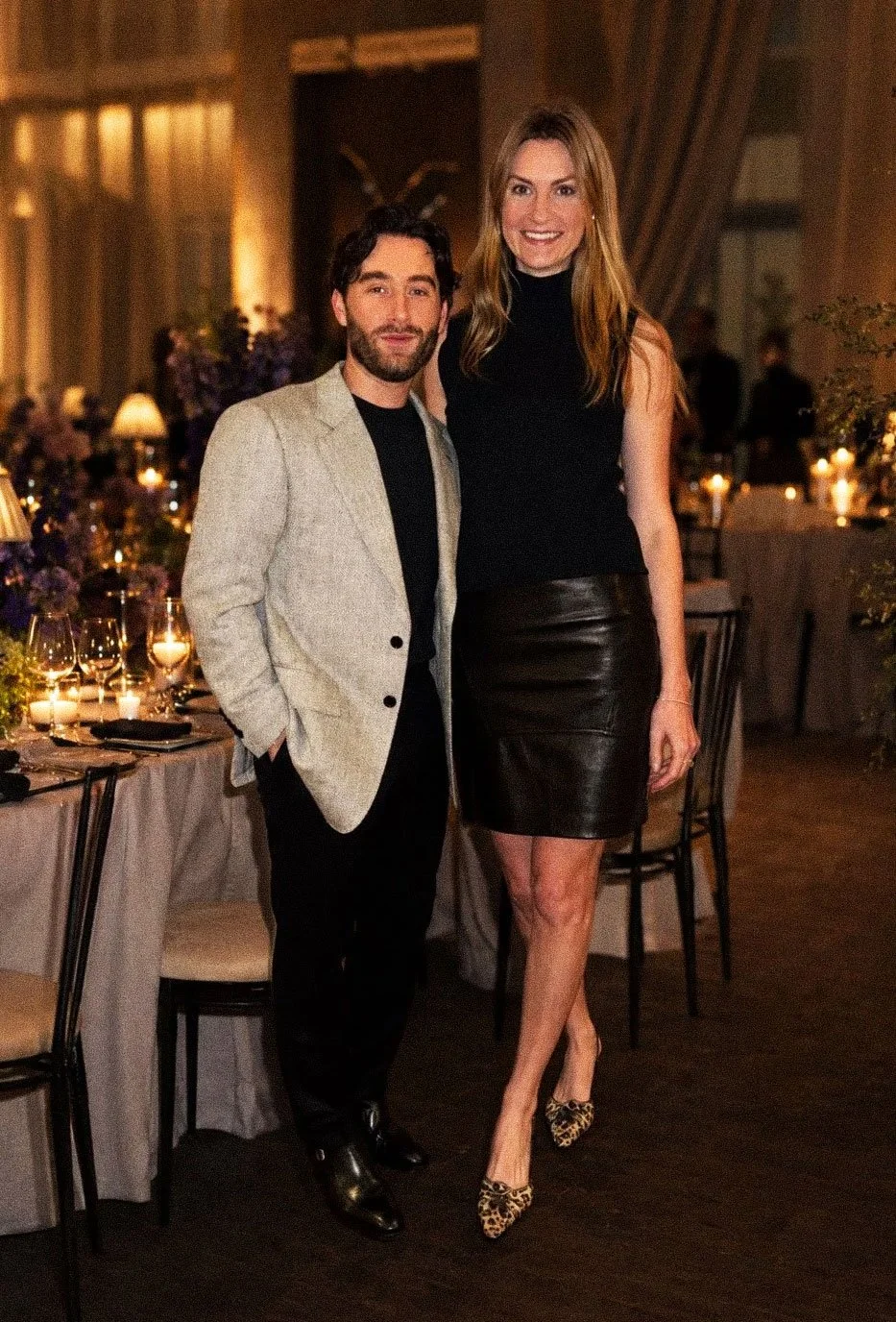 A man and woman stand together at a dinner event, smiling at the camera. The man wears a light gray blazer, black shirt, and black pants. The woman wears a black sleeveless top, a black leather skirt, and leopard print high heels. The background shows a dimly lit room with decorated tables, candles, and flowers.