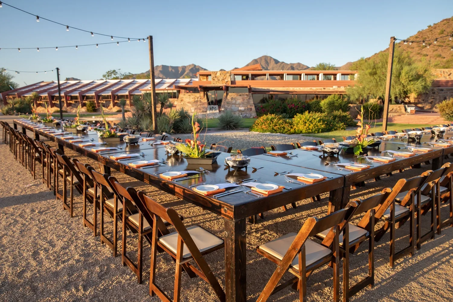Outdoor dining setup with a long wooden table and chairs, set for a meal with plates, napkins, and silverware, in a desert landscape with mountains in the background and string lights overhead.