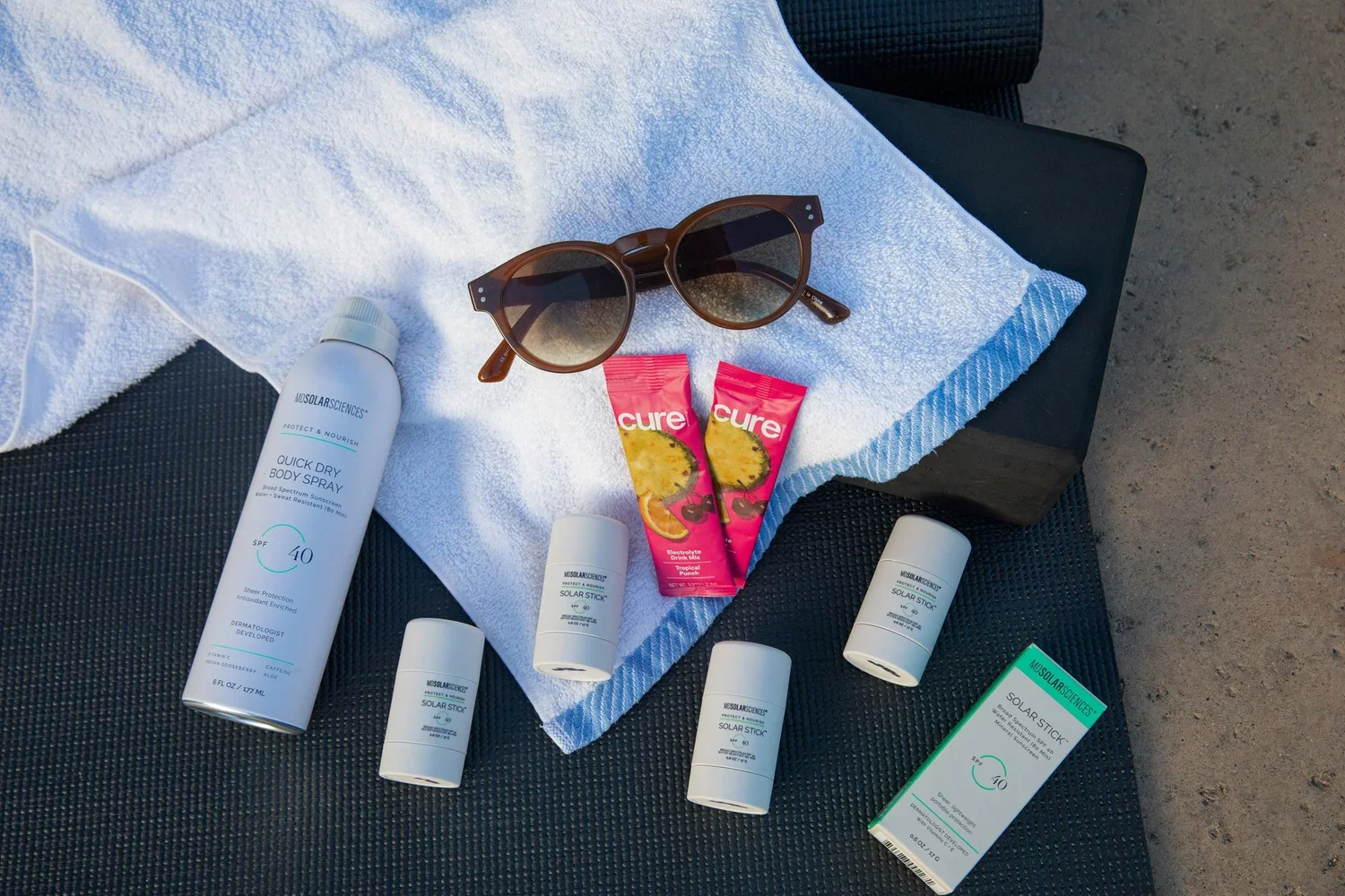 Sun protection products including a large spray bottle, four smaller spray tubes, a box of sunscreen sticks, sunglasses, a white towel, and a packet of Cure electrolyte drinks on a black surface outside.