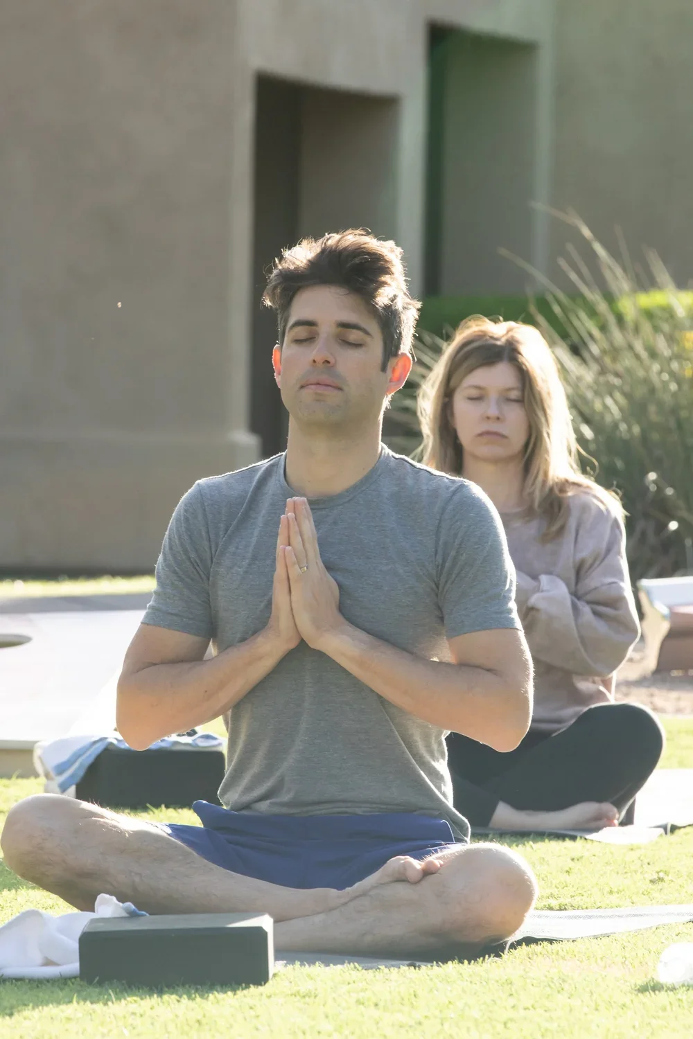 A man and a woman practicing yoga outdoors on the grass, with the man sitting cross-legged in the foreground with eyes closed and hands in a prayer position, and the woman sitting behind him also with eyes closed and hands together.