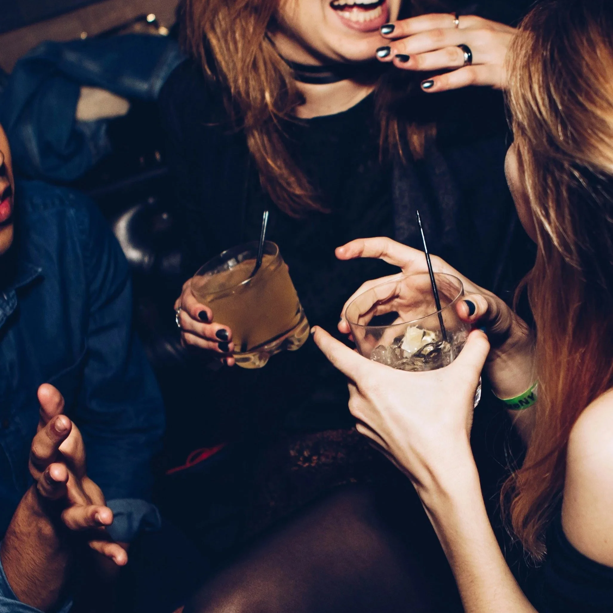 A group of people sharing drinks, with one person holding a glass with ice and a dark beverage. They seem to be at a social gathering or party, and some are wearing rings and jewelry. The focus is on the hands and partial faces.