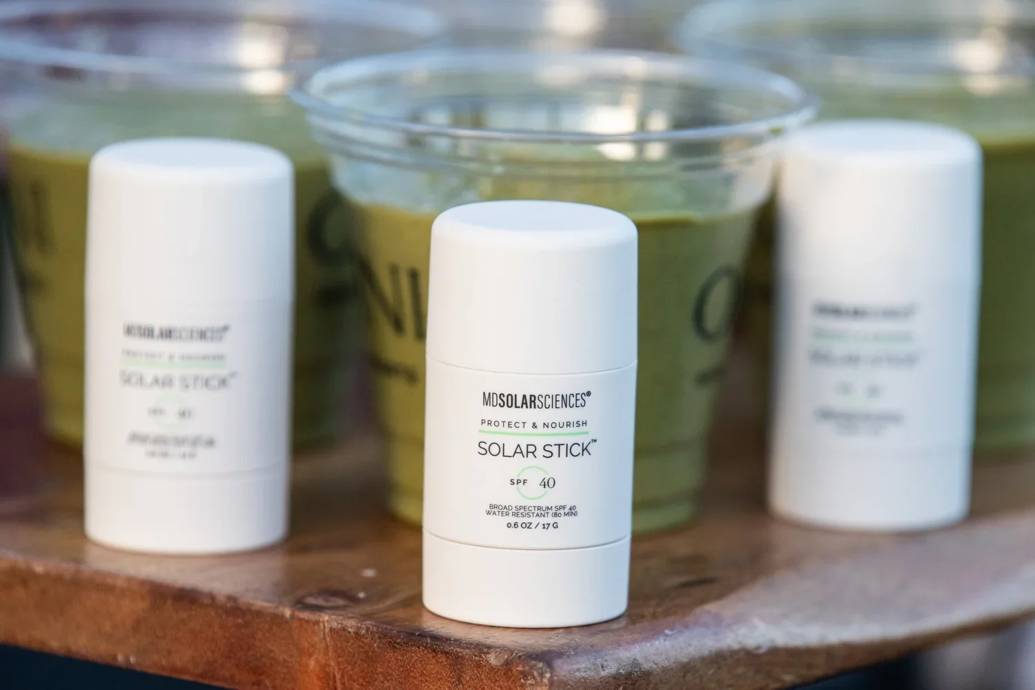 A small white stick container labeled 'MDSOLARSCIENCES Protect & Nourish Solar Stick SPF 40', placed on a wooden surface with blurred green liquid containers in the background.