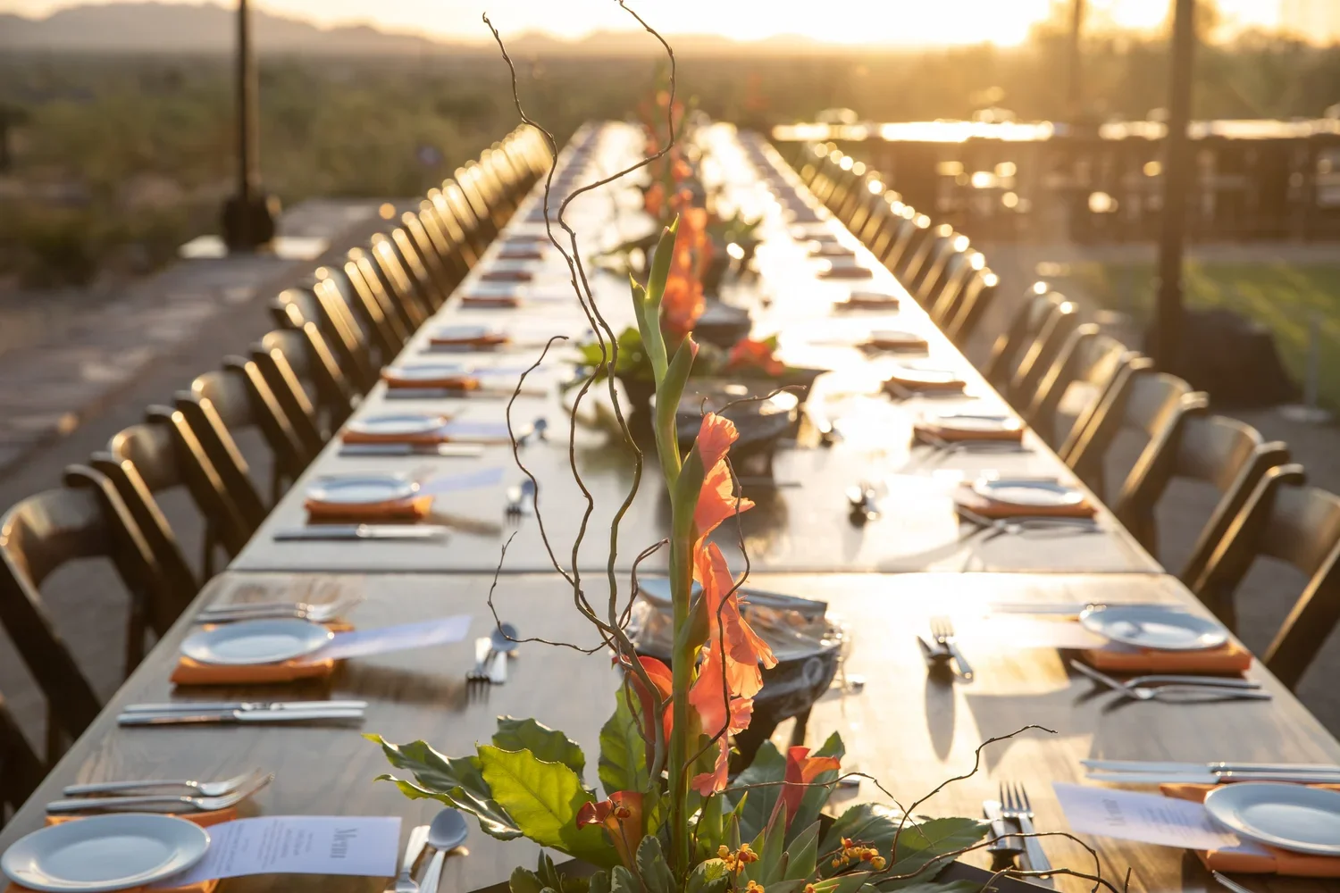 Long outdoor dining table set for a formal event at sunset with floral centerpiece, napkins, plates, and utensils.