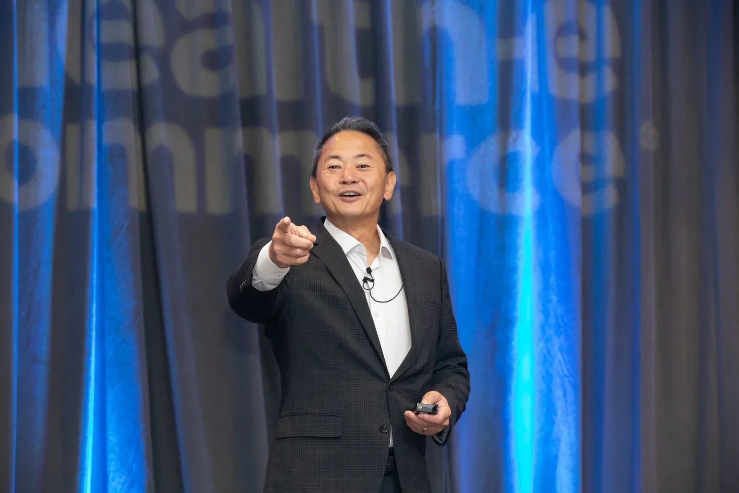 A man in a black suit and white shirt pointing forward while speaking on stage, with blue/gray curtains in the background.