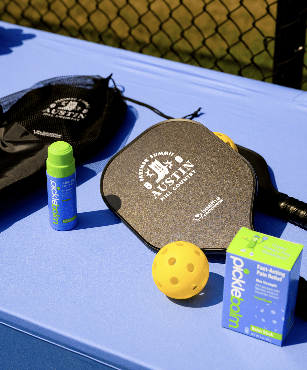 Outdoor tennis scene with safety packing essentials on a blue table, including a yellow tennis ball, sunscreen roll-on, a yellow tennis ball holder, and a box of Pocketbun pain relief balm.