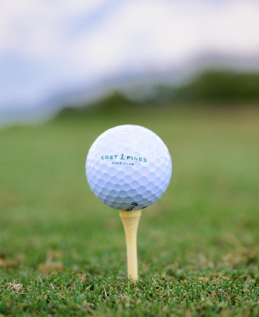 Close-up of a white golf ball with the logo 'Lost Pines Golf Club,' placed on a tee on a golf course grass, with a blurred background of trees and sky.