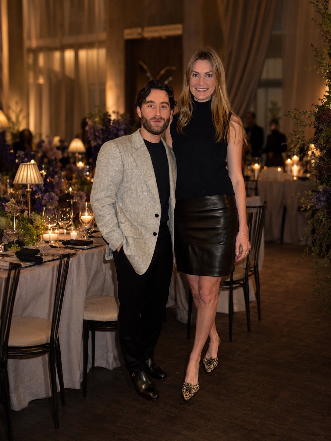 A man and woman standing together at a formal event, with the man in a light gray blazer and black pants, and the woman in a black sleeveless top and leather skirt, in a decorated dinner setting with candles and flowers.