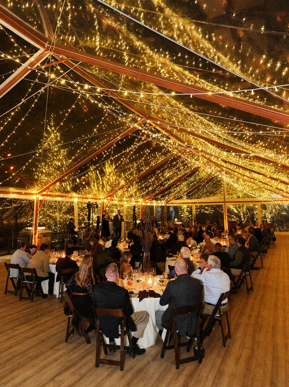 Guests seated at tables under a roof with string lights, at night, with reflections of the lights on the ceiling, during an indoor event or celebration.