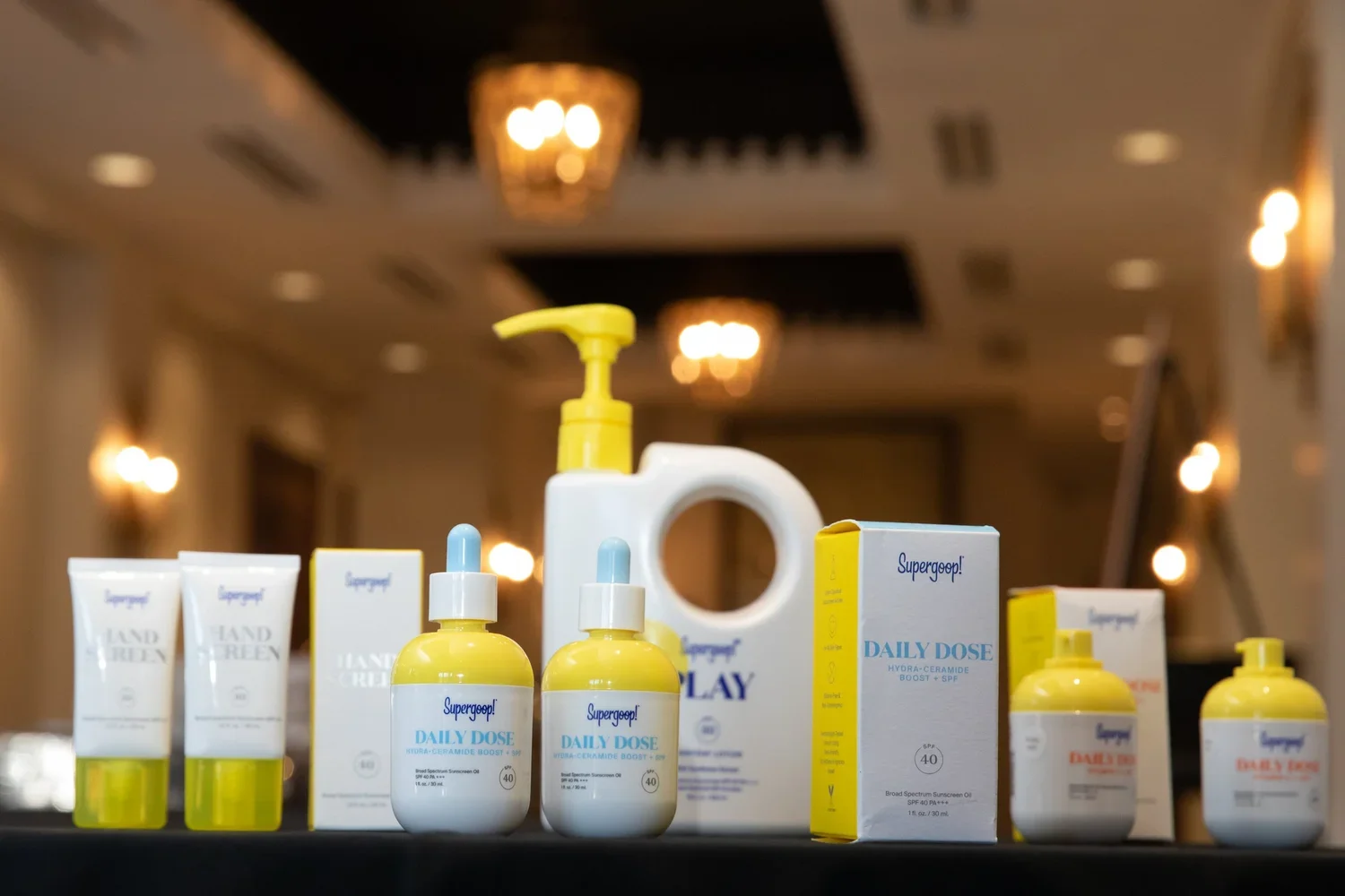Collection of skincare and sunscreen products arranged on a black surface, including bottles and boxes labeled 'Supergoop! Daily Dose' and 'Hand & Body' lotion, with a blurred indoor setting with ceiling lights in the background.