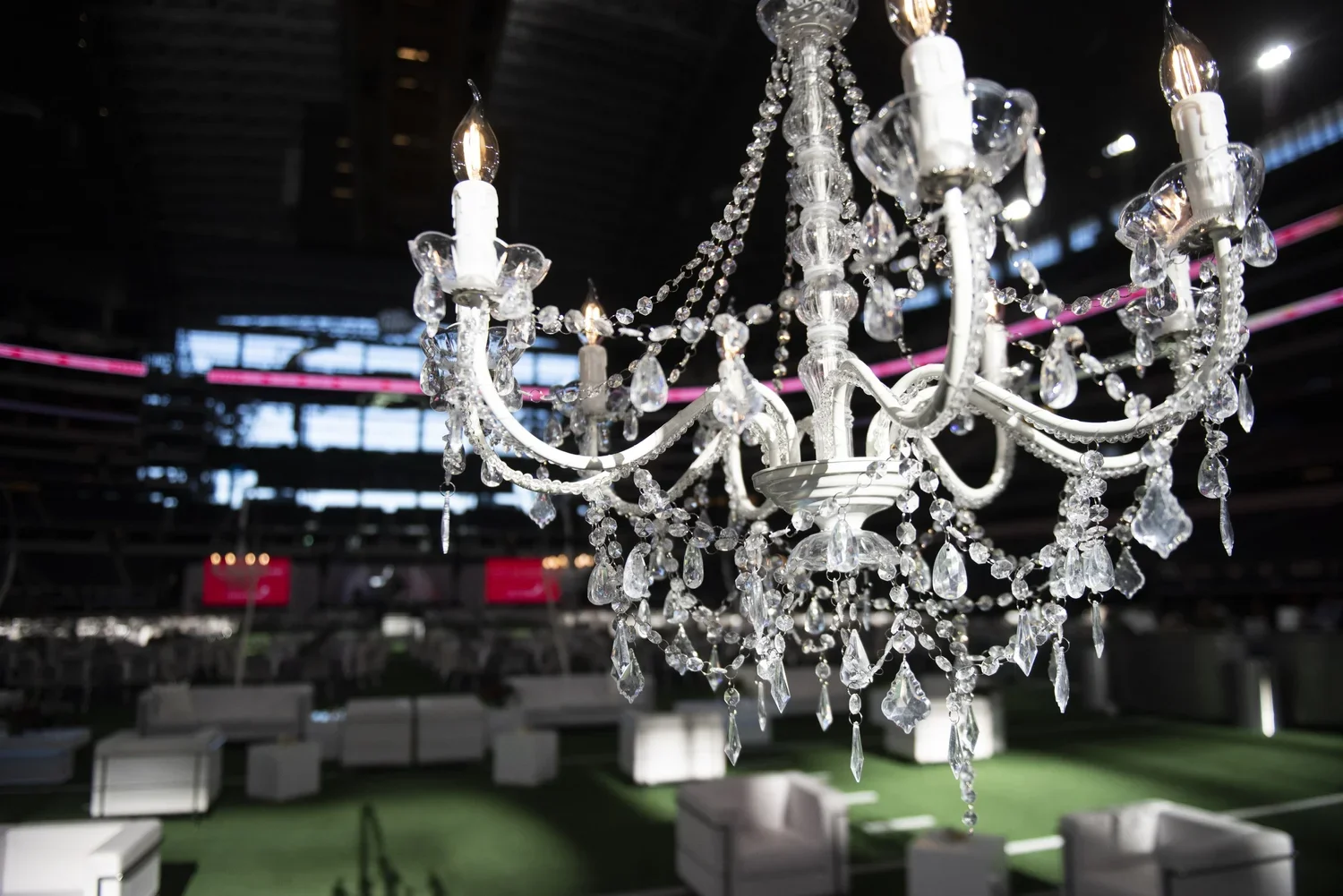 A chandelier with crystal decorations hanging from a ceiling in a large indoor space, possibly a stadium or concert venue, with seating and green flooring below.