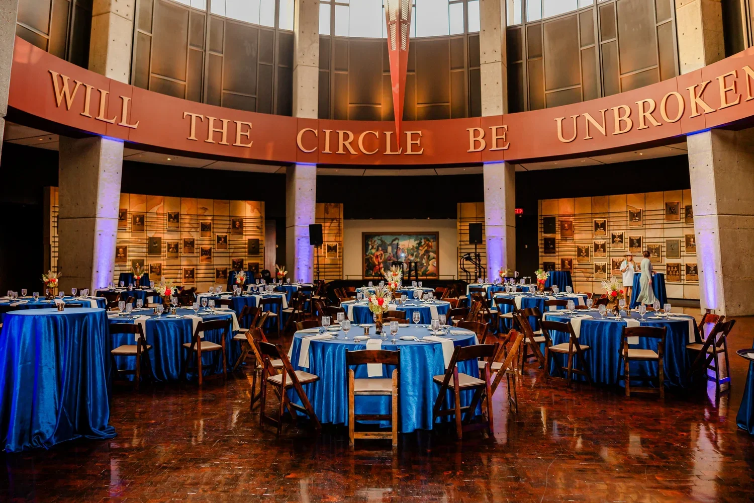 Elegant event space with round tables draped in blue tablecloths, set for a formal dinner with flower centerpieces, inside a modern building with high ceilings and art on the walls. A large orange banner with the words "WILL THE CIRCLE BE UNBROKEN" h