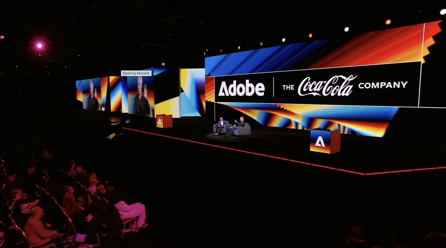 A large conference stage with a digital backdrop displaying the Adobe and Coca-Cola logos, two speakers sitting in chairs, and an audience watching.